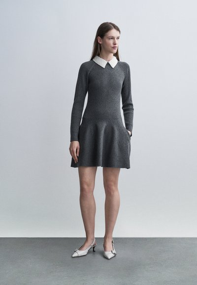 Femme debout portant une robe en maille côtelée gris foncé avec col pointu blanc et escarpins slingback gris clair à petit talon, mains détendues.