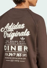 Hnědá mikina s potiskem "Adidas Originals" a grafickým designem na zadní straně, s kulatým výstřihem a měkkou texturou.