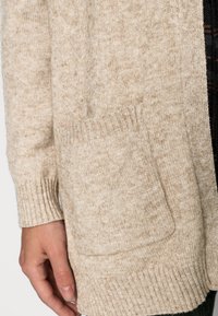 Manica di un cardigan lavorato a maglia beige con polsino a coste e tasca frontale, che copre parzialmente una mano appoggiata accanto al corpo.