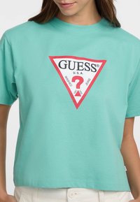Turkio spalvos medvilnės marškinėliai su raudonu trikampiu logotipu, kuriame yra „GUESS U.S.A.“ ir klausimo ženklas centre. Trumpomis rankovėmis, laisvas kirpimas.