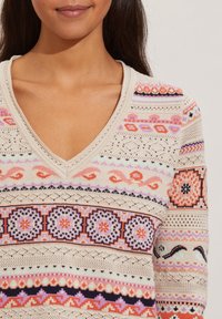 Pull en tricot à col en V avec motifs géométriques et floraux multicolores sur fond crème. Tissu texturé avec accents en maille ajourée.