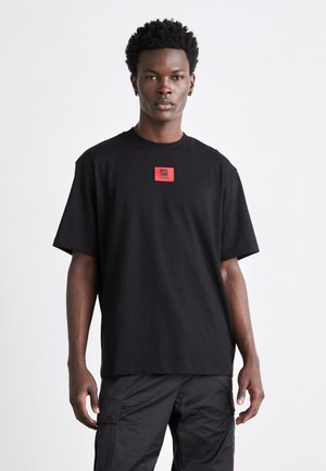T-shirt en coton noir avec un col rond et des manches courtes. Dispose d'un patch logo rouge carré sur la poitrine. Coupe droite, texture minimale.
