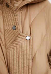 Beige gewatteerde pufferjack met een diagonale stikpatroon en twee drukknoopjes. Glad, waterafstotend stof met een hoge kraag.