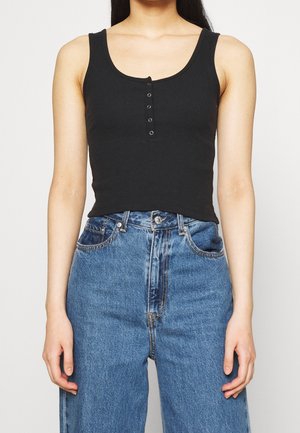 Kobieta ubrana w czarny, bezrękawnikowy, prążkowany, krótki top na zatrzaski oraz wysokim stanem niebieskie dżinsy na jednolitym tle.