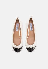 Baltās baleta kurpes ar melniem lakotiem purngalu vāciņiem, maziem lokiem un brūnām ieliktņām, uz kuriem rakstīts "Steve Madden", rādītas no augšas uz balta fona.