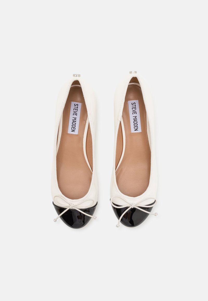 Ballerines blanches avec des bouts en vernis noirs, petites accents en forme de nœud, et semelles beige étiquetées "Steve Madden", vues de dessus sur un fond blanc.