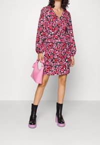 Květinové šaty s růžovými a černými odstíny, véčkový výstřih, dlouhé nabírané rukávy; spárováno s malou růžovou kabelkou a černými platformovými kotníkovými botami.