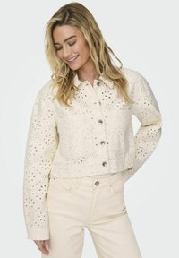 Veste courte crème en matière douce avec des motifs floraux découpés au laser, boutonnée à l'avant et dotée de deux poches poitrine ; assortie à un pantalon clair.