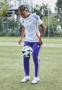 Adidas sportkläder har en vit skjorta med orange detaljer, lila byxor med tre ränder och vita fotbollsskor, medan en fotboll balanseras.