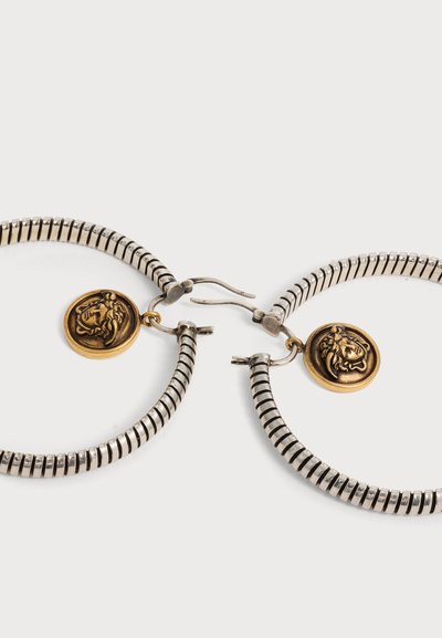 Boucles d’oreilles créoles argentées à texture rainurée, chacune ornée d’un petit médaillon doré embossé d’une tête de lion.