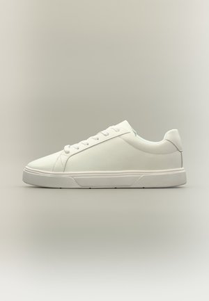 Witte lage sneaker met veters, eenvoudig ontwerp en een gestructureerde rubberen zool op een effen neutrale achtergrond.