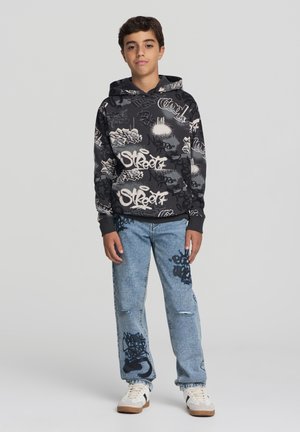 Felpa nera con motivo graffiti e tasca a marsupio, abbinata a jeans chiari strappati con accenti blu scuro e sneakers bianche.