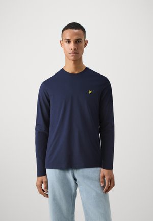 Tricou cu mânecă lungă de culoare bleumarin, confecționat dintr-un material neted, cu guler rotund și un mic logo galben pe piept, asortat cu blugi de culoare bleu deschis.
