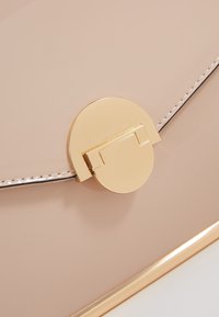 Světle růžová kabelka z lakované kůže se zlatým kulatým zapínáním, která má hladkou texturu a tenké ozdobné šití podél okrajů.