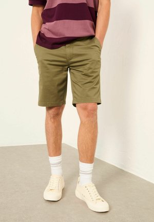 Mann trägt olivgrüne Shorts, weiße Socken, cremefarbene Sneaker und ein bordeauxgestreiftes Hemd, steht mit den Händen in den Taschen auf grauem Boden.