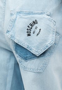 Ανοιχτό μπλε τζιν παντελόνι με δύο πίσω τσέπες, η μία επάλληλη της άλλης, με την επιγραφή "MOSCHINO 86-23 JEANS" σε μαύρο στην πάνω τσέπη.