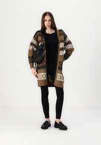 Cárdigan de punto marrón con patrones geométricos en blanco y negro, llevado sobre una camiseta negra y jeans negros, combinado con un bolso y zapatos negros.