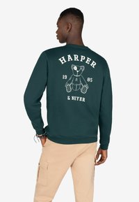 Sudadera verde de cuello redondo con un gráfico de oso de peluche en la parte posterior, junto con el texto "HARPER 1985 & NEYER" en blanco. Tejido suave con puños acanalados.