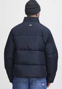 Navy puffer jacket with a high collar, quilted design, and logo tag on the back. Paired with blue jeans showing pockets and stitching details.