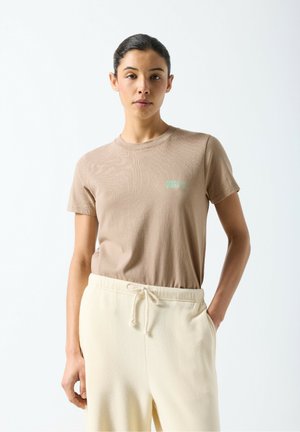 Jasnobrązowy bawełniany T-shirt z okrągłym dekoltem i krótkimi rękawami, z małym zielonym logo na piersi. Kremowe spodnie ze ściągaczem.