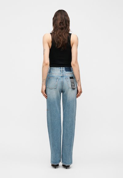 Γυναίκα με μακριά καστανά μαλλιά στέκεται πλάτη, φοράει μαύρο αμάνικο τοπ, ανοιχτό μπλε τζιν με φαρδιά πόδια με τρουκς στις τσέπες και διακοσμητική επιφάνεια, μαύρες γόβες.