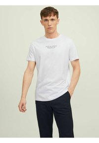 Bílý bavlněný tričko s kulatým výstřihem, krátkými rukávy a černým textovým logem "JACK & JONES PREMIUM" na hrudi. Model má na sobě tmavé kalhoty.