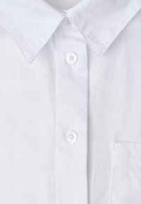Chemise blanche boutonnée à l'avant avec col pointu, deux boutons transparents fermés, et une vue partielle d'une poche poitrine sur un tissu texturé.