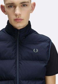 Giovane uomo con capelli corti che indossa un gilet trapuntato blu navy abbottonato sopra una maglietta nera a maniche corte, con un logo a corona d'alloro sul petto.