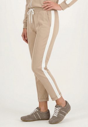 Beige joggerbukser med hvide sidestriber og op rullede kanter, båret med grå afslappede sneakers med snørebånd på en person, der står.