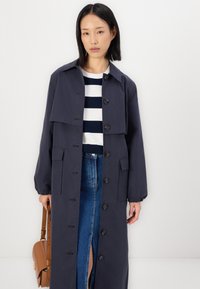 Cappotto lungo blu navy con colletto, bottoni frontali e maniche elastiche; indossato sopra una maglietta a righe e una gonna blu. Presenta due tasche frontali.