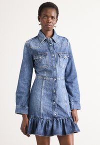 Vestido de ganga com um tom de azul claro, frente com botões, grandes bolsos no peito e bainha com volantes. Apresenta um colarinho pontiagudo e mangas longas.