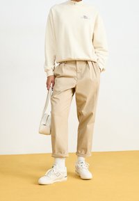 Krämfärgad zip-up sweatshirt med ljusbeige byxor och vita sneakers. Modellen håller en liten vit handväska. Gul bakgrund.