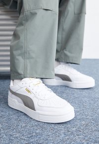 Vita lädersneakers med grå detaljer och gulddekor, med en tjock sula och perforeringar på tån, som bärs med gröna byxor.