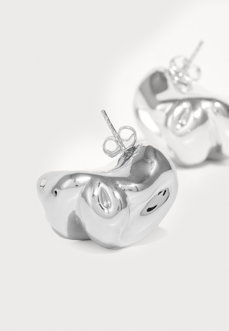 Ασημί μεταλλικά σκουλαρίκια με γυαλιστερή φινέτσα, σε αφηρημένο σχήμα που μοιάζει με μια φόρμα με μικρή στήριξη.