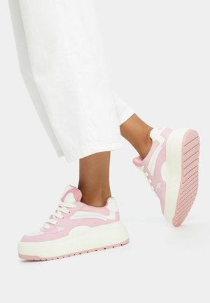 Persona que lleva pantalones blancos y zapatillas plataforma gruesas rosas y blancas, con un pie levantado mostrando la suela del zapato.