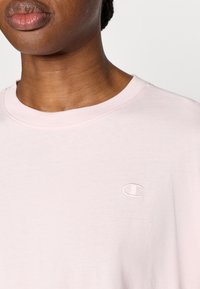 Ροζ ανοιχτό μπλουζάκι με στρογγυλή λαιμόκοψη και κεντημένο λογότυπο στο στήθος. Το ύφασμα φαίνεται μαλακό με απαλό υφή.