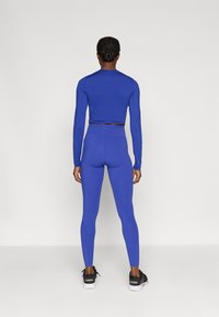 Plavi dug rukav uski crop top koji je uparen s visokim plavim tajicama. Odjeća ima glatku teksturu i prilagođen dizajn. Vidi se crne atletske tenisice.