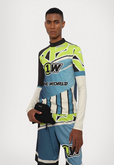 Langærmet grafisk rash guard og shorts i blå, grøn, sort og hvid med geometriske mønstre og et logo. Modellen holder et sort tilbehør.