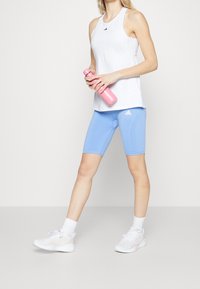 Biały sportowy top na ramiączkach i jasnoniebieskie szorty rowerowe, w połączeniu z białymi trampkami. Trzyma różową butelkę na wodę. Z logo Adidas.