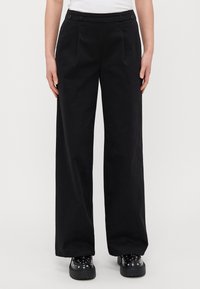 Black wide-leg trousers with pleats, featuring a smooth texture and two button accents on the waistband. Paired with chunky black footwear.
