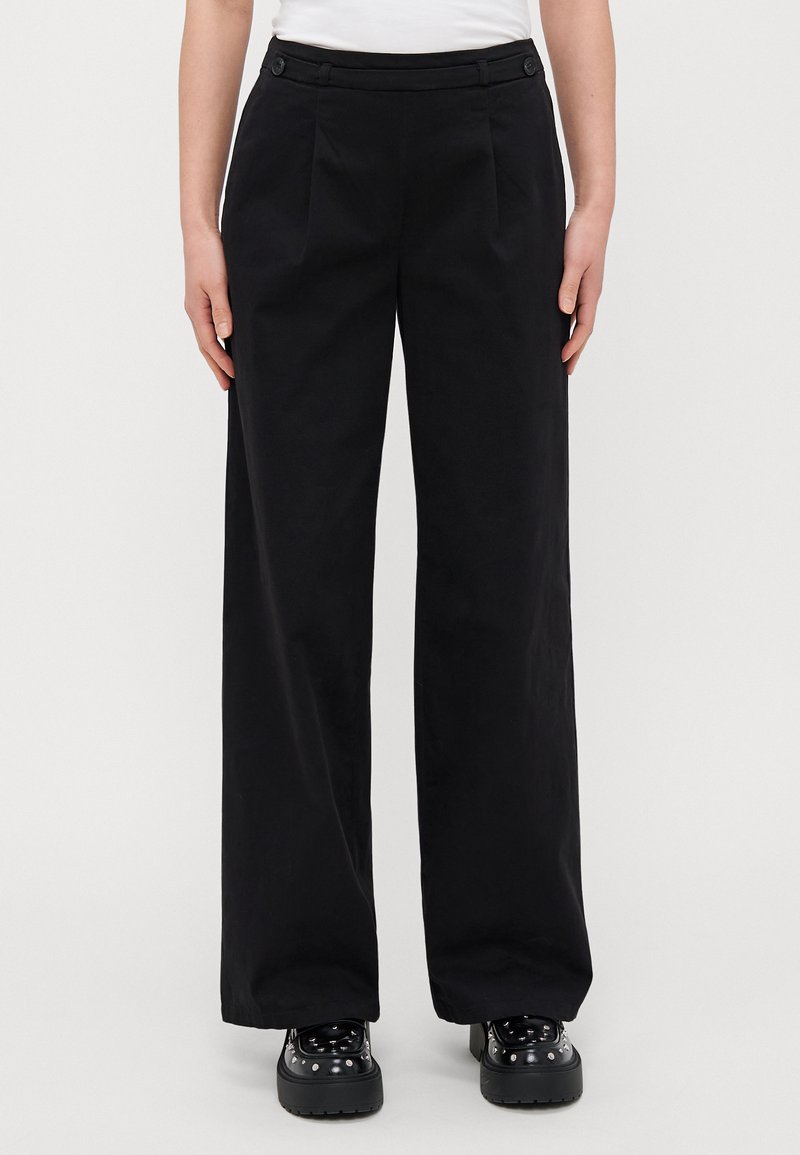 Black wide-leg trousers with pleats, featuring a smooth texture and two button accents on the waistband. Paired with chunky black footwear.