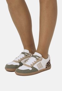 Piedi indossanti scarpe da ginnastica bianche, beige e verdi con dettagli leopardati sul tallone, in piedi su uno sfondo bianco.