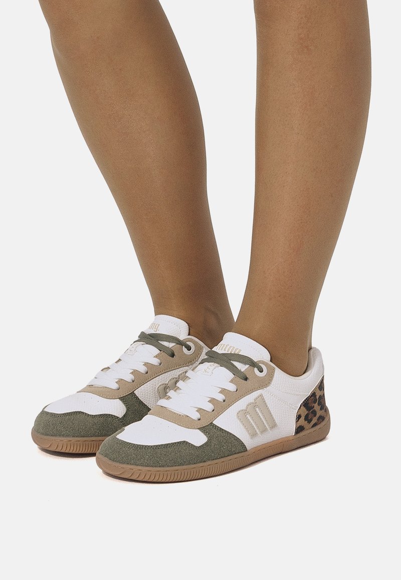 Piedi indossanti scarpe da ginnastica bianche, beige e verdi con dettagli leopardati sul tallone, in piedi su uno sfondo bianco.