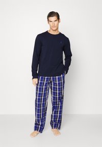 Navy blauwe lange mouw shirt met een klein logo en blauwe geruite katoenen pyjamabroek met witte en zwarte accenten. Model staat blootsvoets tegen een effen achtergrond.