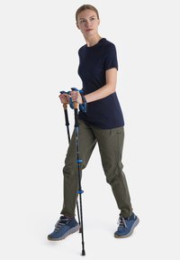 Turista ve tmavě modrém tričku a olivově zelených kalhotách, držící černé a modré trekingové hole, obutý v modrých turistických botách s oranžovými podrážkami.