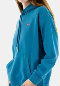 Tyrkysová mikina s kapucí na šňůrku a přední kapsou. Na hrudi má malé červené logo Levi's a vroubkované manžety. Hladká textura.