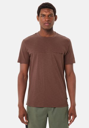 Uomo in piedi di fronte indossa una maglietta marrone a maniche corte e pantaloni verdi davanti a uno sfondo bianco uniforme.