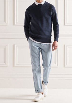 Maglione strutturato blu navy con maniche raglan sopra una camicia bianca con colletto, abbinato a pantaloni azzurri risvoltati e sneakers bianche.