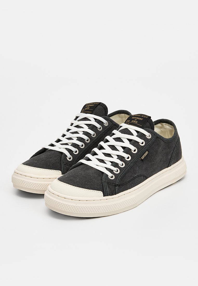 Zapatillas de lona negras con cordones blancos, con suela de goma y una puntera color crema. Etiqueta con el logo en la lengua, diseño simple.