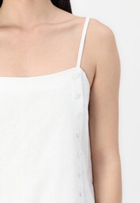 White linen camisole with thin straps and a square neckline, featuring a row of round fabric-covered buttons along the left side.